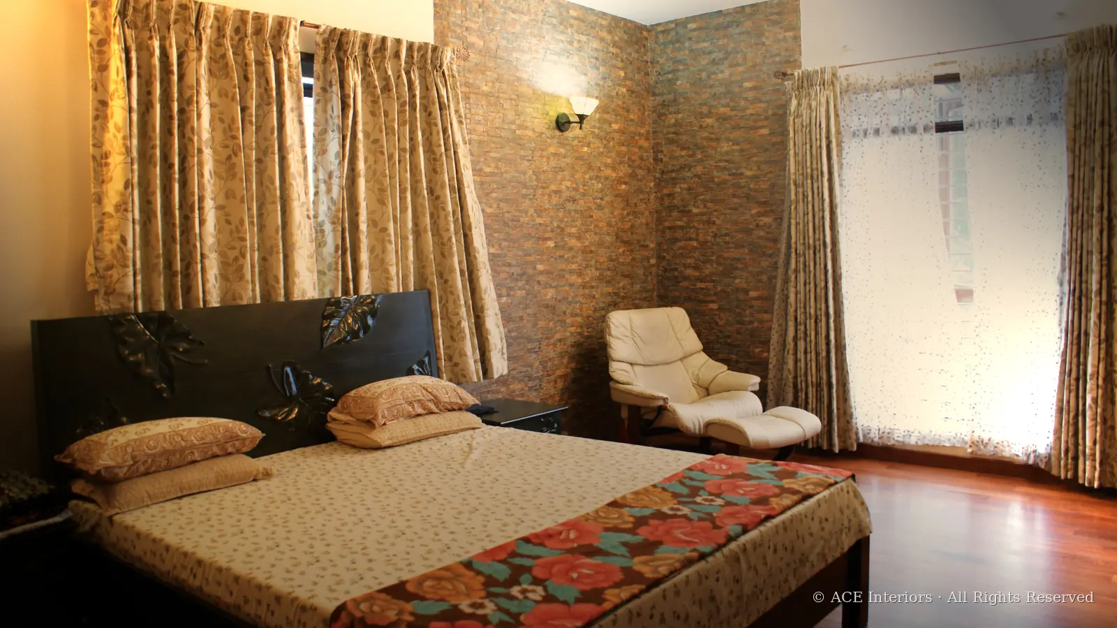 A warm ACE Interiors bedroom with draped windows, a stone feature wall and a carved wood bed